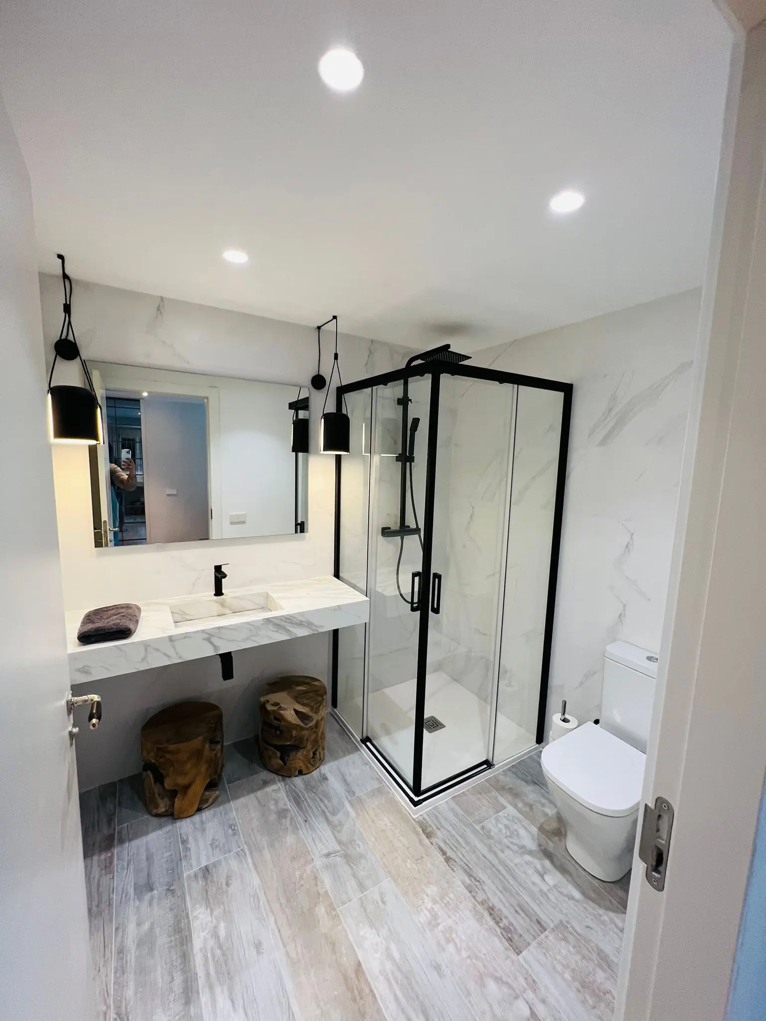 Baño moderno reformado con ducha de vidrio, lavabo de mármol y taburetes de madera.