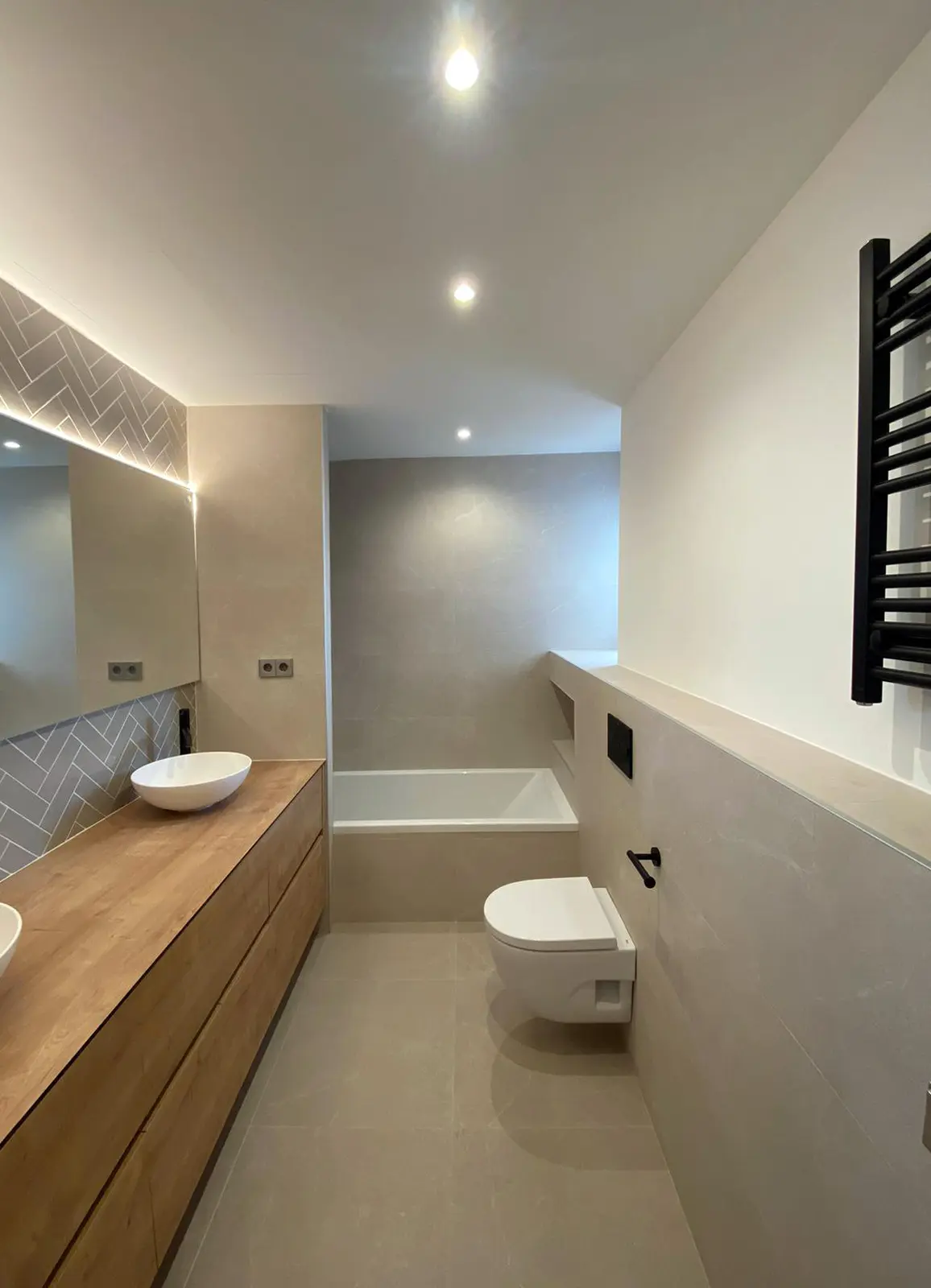 Este proyecto de reforma destaca una cocina moderna y minimalista con una isla central que incluye iluminación LED en la base, creando un efecto visual elegante. Los acabados en madera natural y las lámparas colgantes de diseño aportan calidez al espacio, mientras que los taburetes combinan madera y blanco para un look sofisticado. La distribución abierta y los detalles cuidadosamente seleccionados hacen de esta cocina un espacio funcional y acogedor, perfecto para la vida diaria y el entretenimiento.