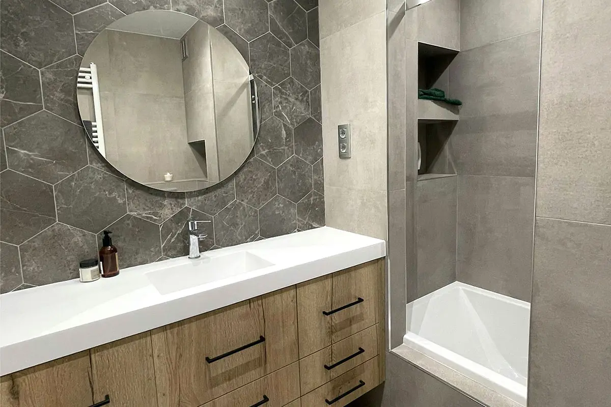 Baño moderno reformado con lavabo de madera, espejo redondo y ducha de azulejos grises.